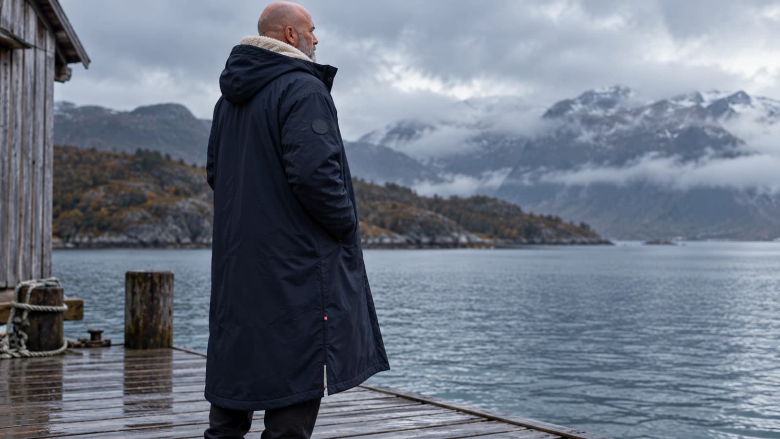 Person i Comfyrobe utendørs ved en skandinavisk fjord i gråvær, der både det matte HydroGuard-ytterstoffet og det lyse SherpaCore-innerfôret i hetten er synlig.