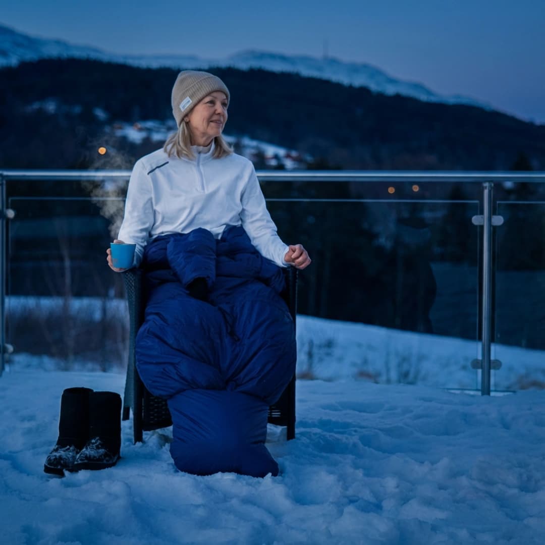 En kvinne iført Utekos-plagg sitter på terrassen i snørike omgivelser.