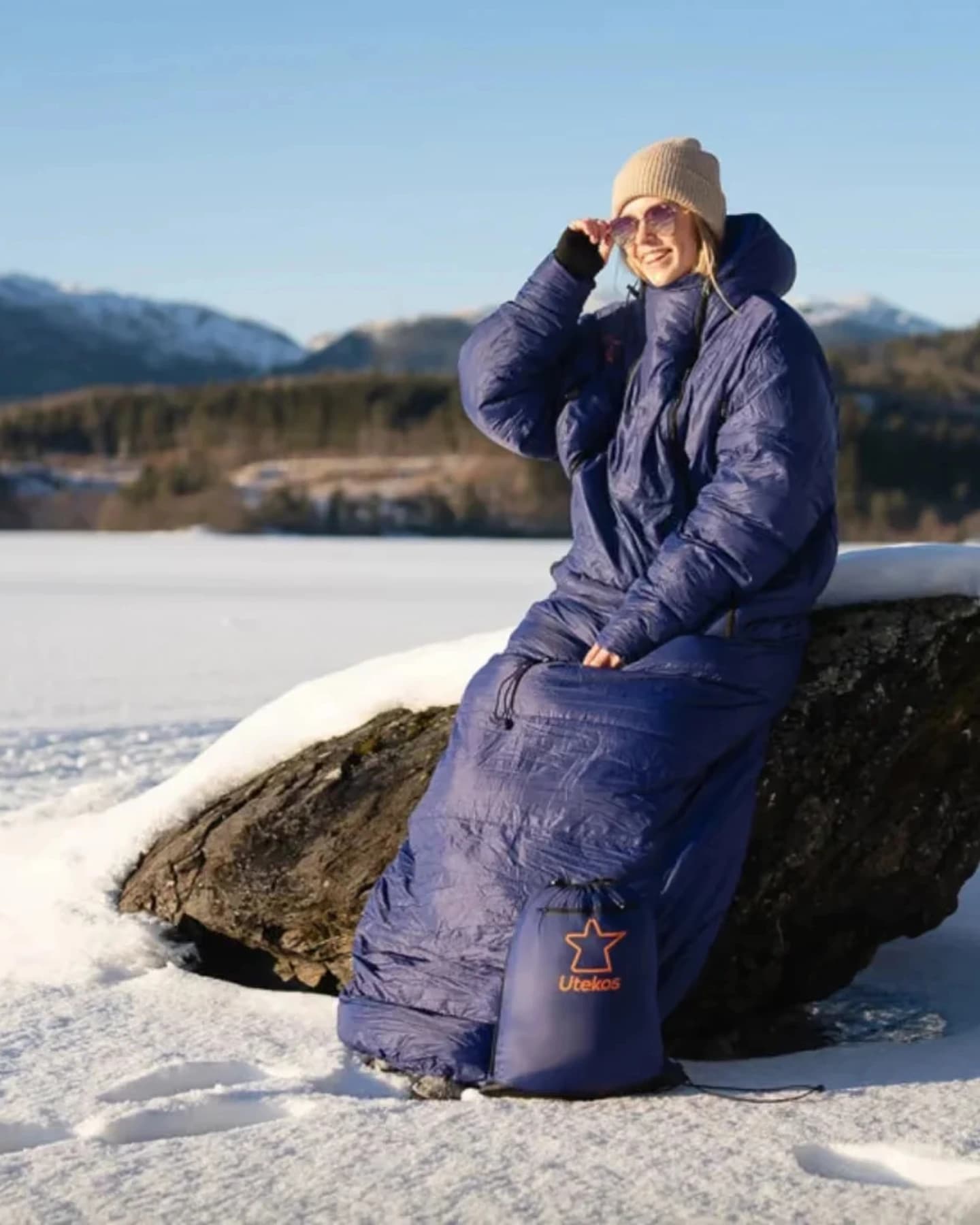 Kvinne i et blått Utekos-plagg på en snødekt stein med fjell i bakgrunnen